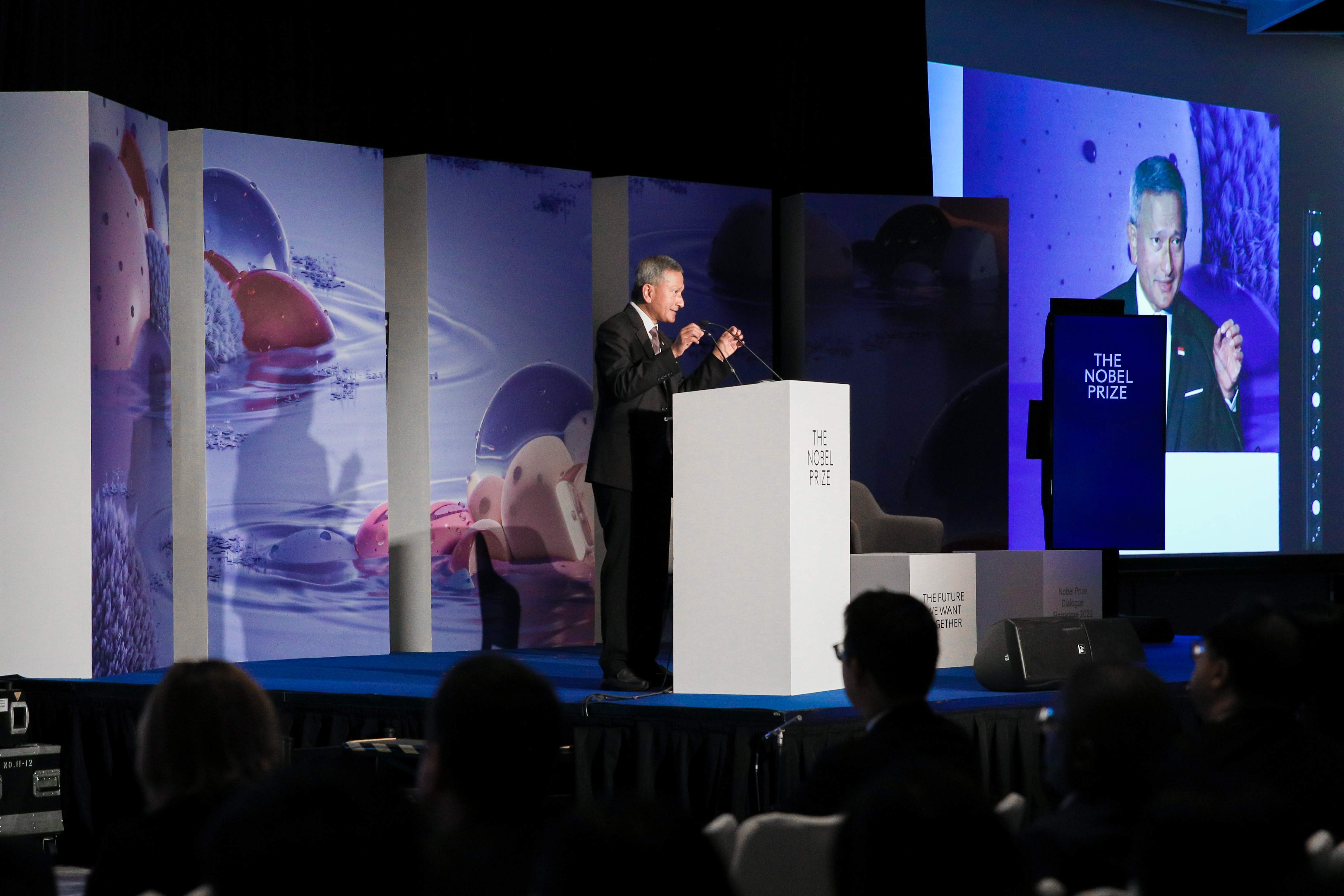 Man speaking at "The Nobel Prize" event with screen showing the same, audience in foreground.